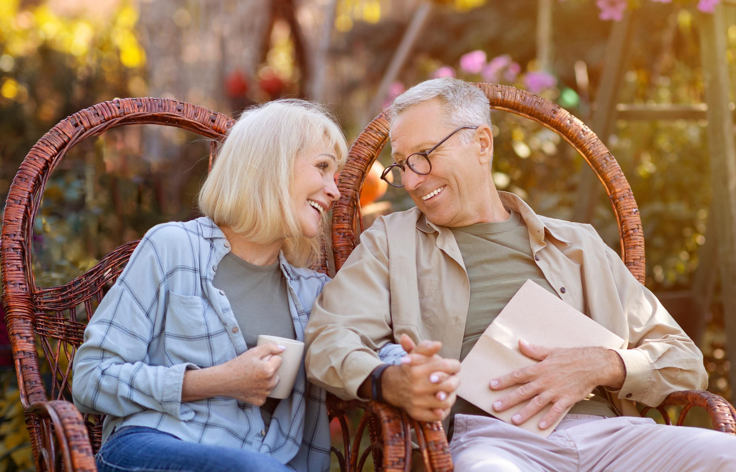 Smiling older couple sitting in wicker chairs outdoors, enjoying their Minnesota retirement. The woman holds a mug, and the man holds a closed book. They lean toward each other, appearing happy and relaxed in a sunlit, garden-like setting.