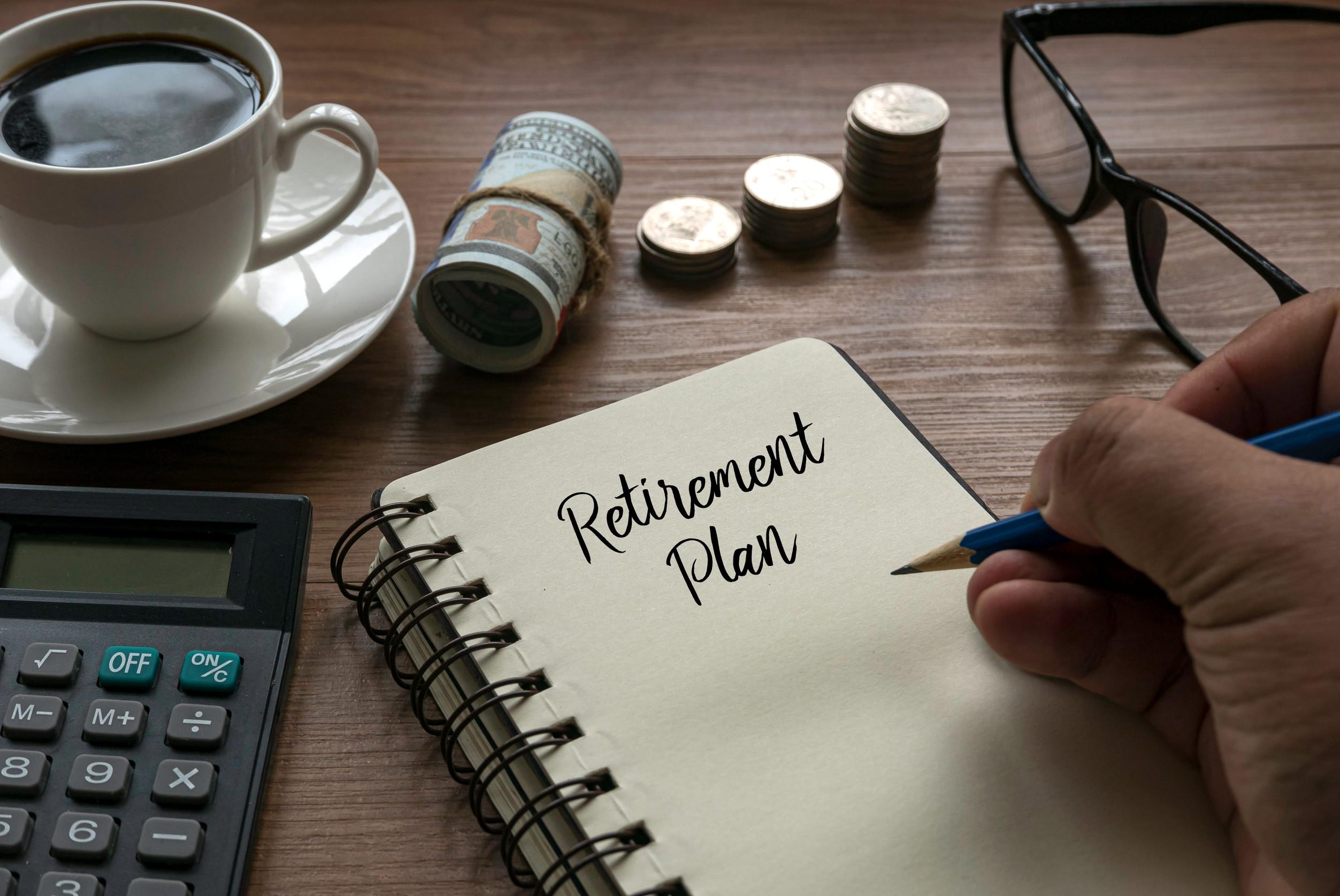 A hand writes “Retirement Plan” in a notebook on a desk with a calculator, cup of coffee, rolled dollar bills, stacked coins, and eyeglasses nearby—perfect inspiration for anyone considering when to start retirement or thinking about effective retirement planning.