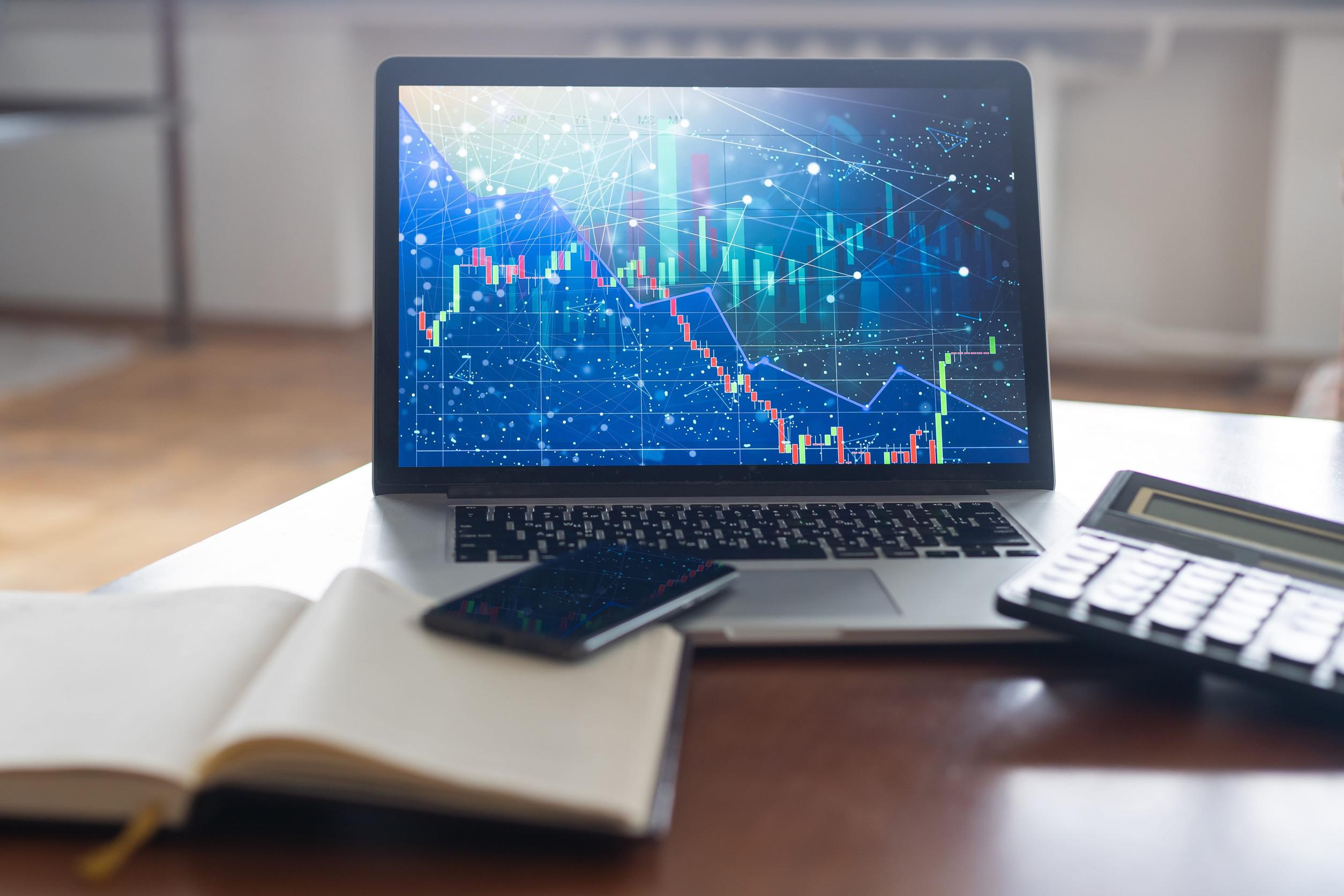 An open laptop displays a digital financial chart with graphs, a notebook, smartphone, and calculator on a desk, suggesting ongoing financial analysis or trading work in progress—ideal for investment management or thorough portfolio reviews.