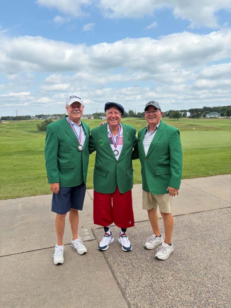 Three men wearing green jackets, medals, and hats stand smiling on a golf course with cloudy skies in the background. One man is wearing red shorts while the others wear khaki and navy shorts.