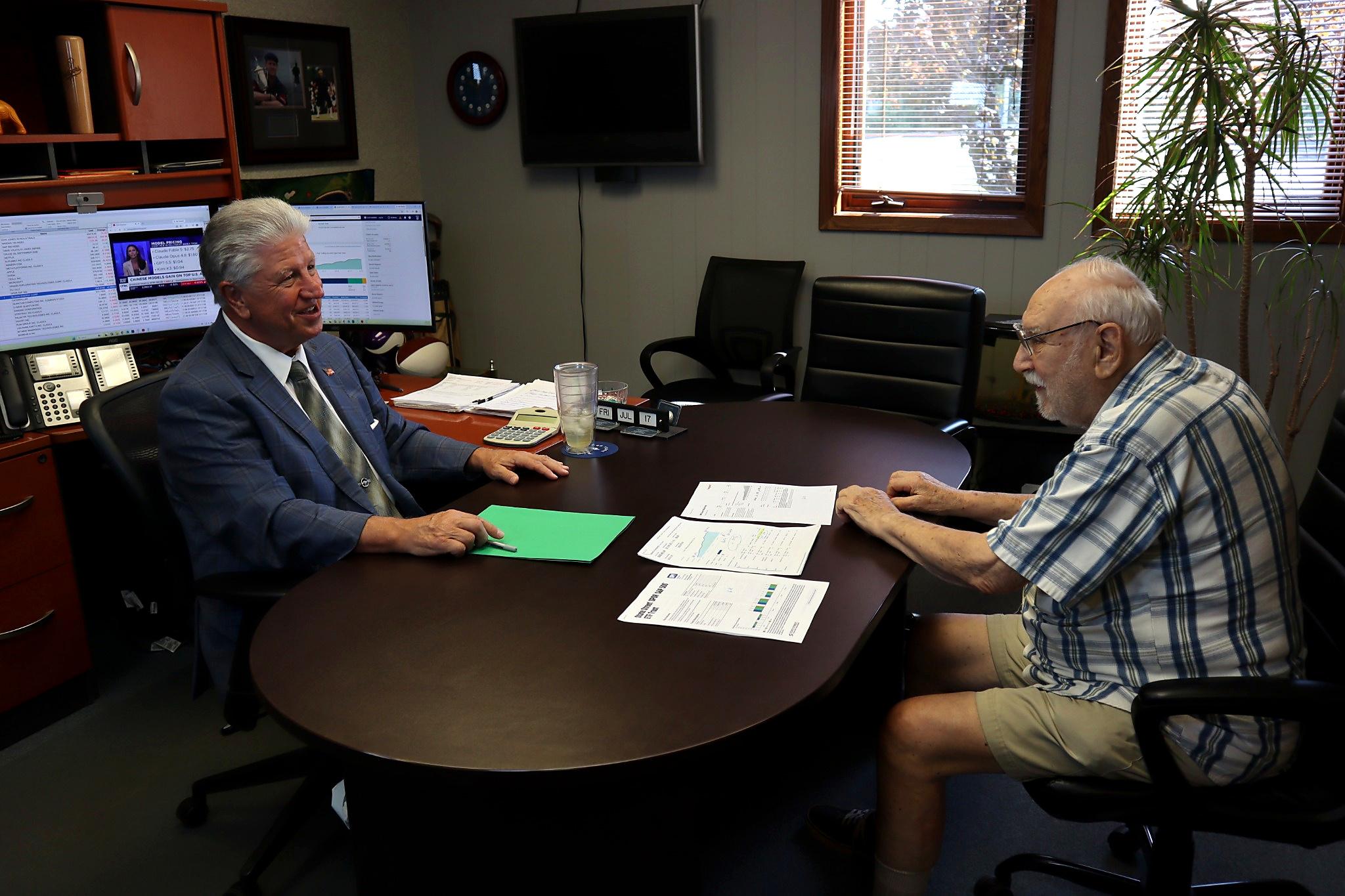 An older man in casual clothes sits across a round table from a man in a suit. In this modern office setting that blends the comfort of a business home, they are discussing papers with charts, emphasizing collaboration and professionalism.