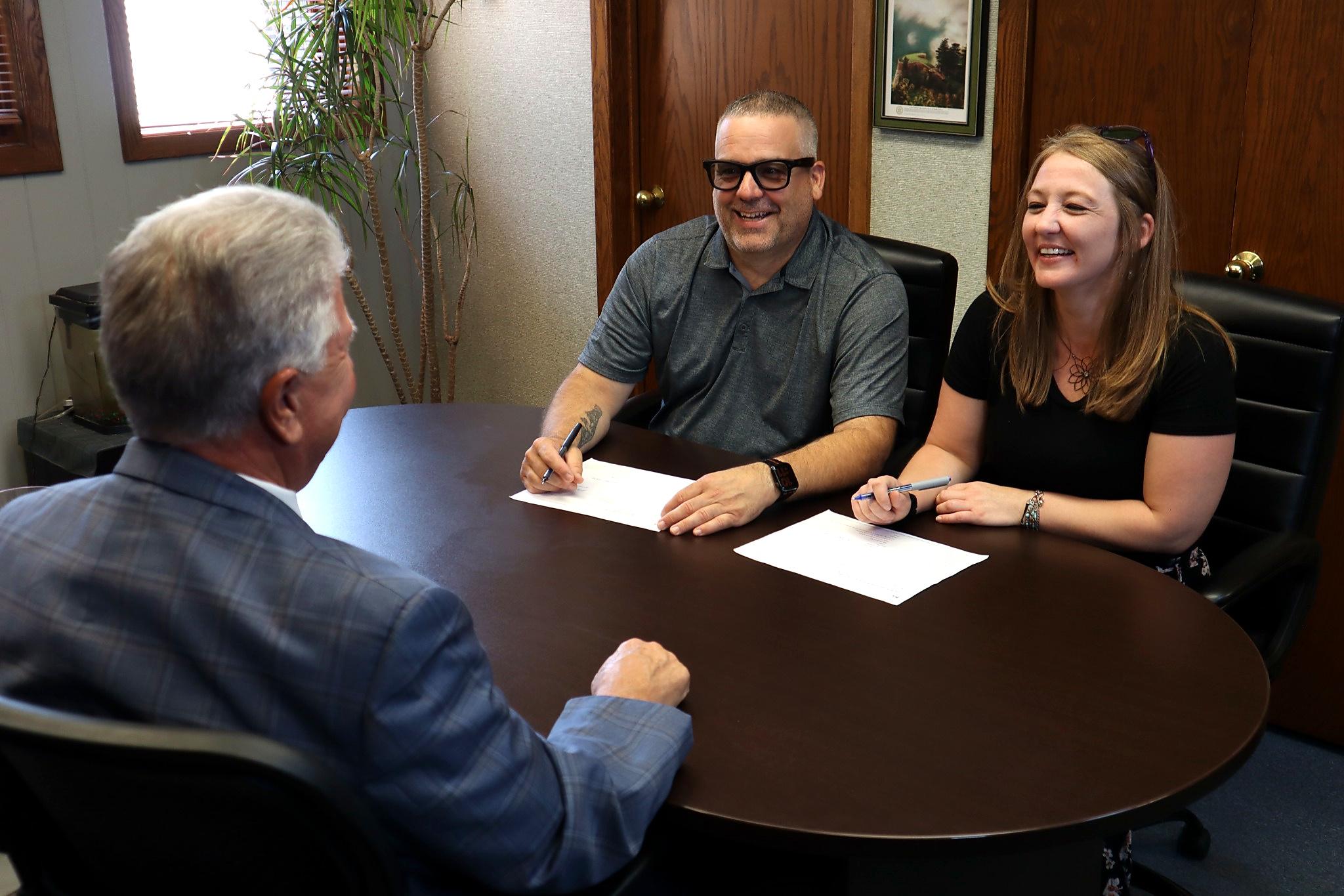 A man and a woman smile while signing documents at a round table with another man in a suit across from them. There are papers and pens on the table in an office setting, suggesting an important moment at their Business Home.