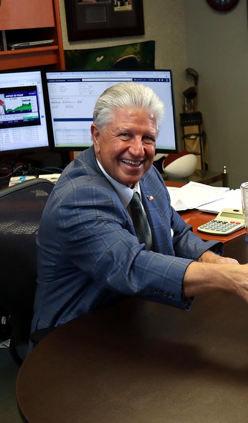A man in a blue suit and a woman with long hair shake hands and smile across a desk covered with papers, while another man sits beside her in what appears to be a modern Business Home office. Computer monitors and office equipment are visible in the background.