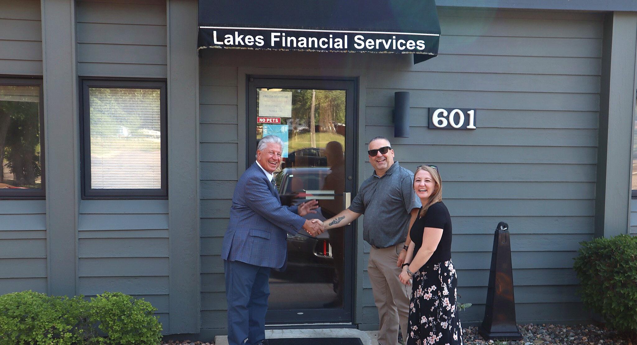 Three people smile outside a building labeled “Lakes Financial Services,” which serves as their trusted Business Home. Two men are shaking hands, and a woman stands beside them. The building is gray with a glass door and the number 601 is visible.