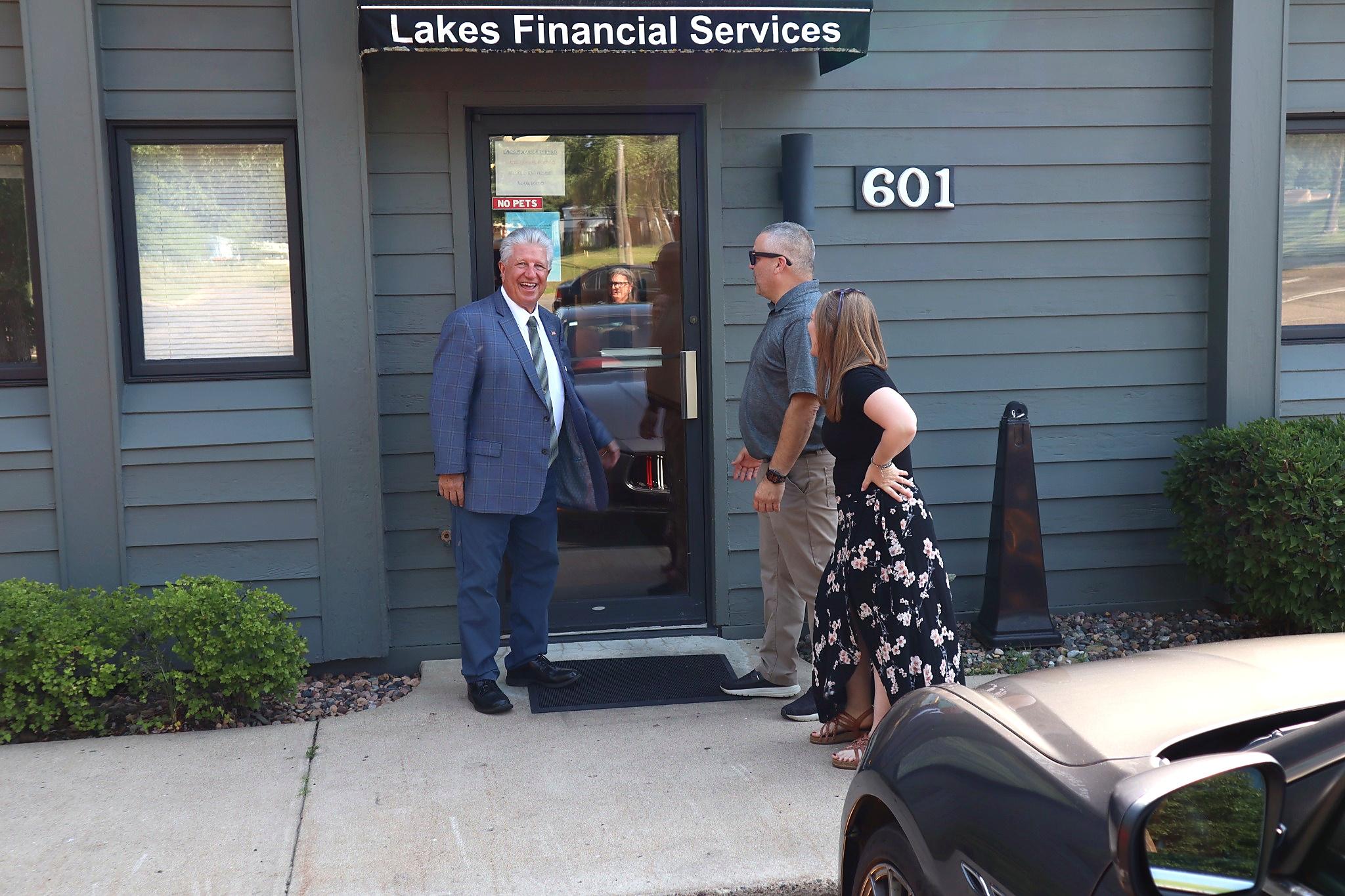 Three people stand talking outside the entrance to Lakes Financial Services, building number 601. One man is wearing a suit, another is in casual clothes, and a woman wears a floral dress—demonstrating the diverse clientele that might find their Business Home here. A car is partially visible in the foreground.
