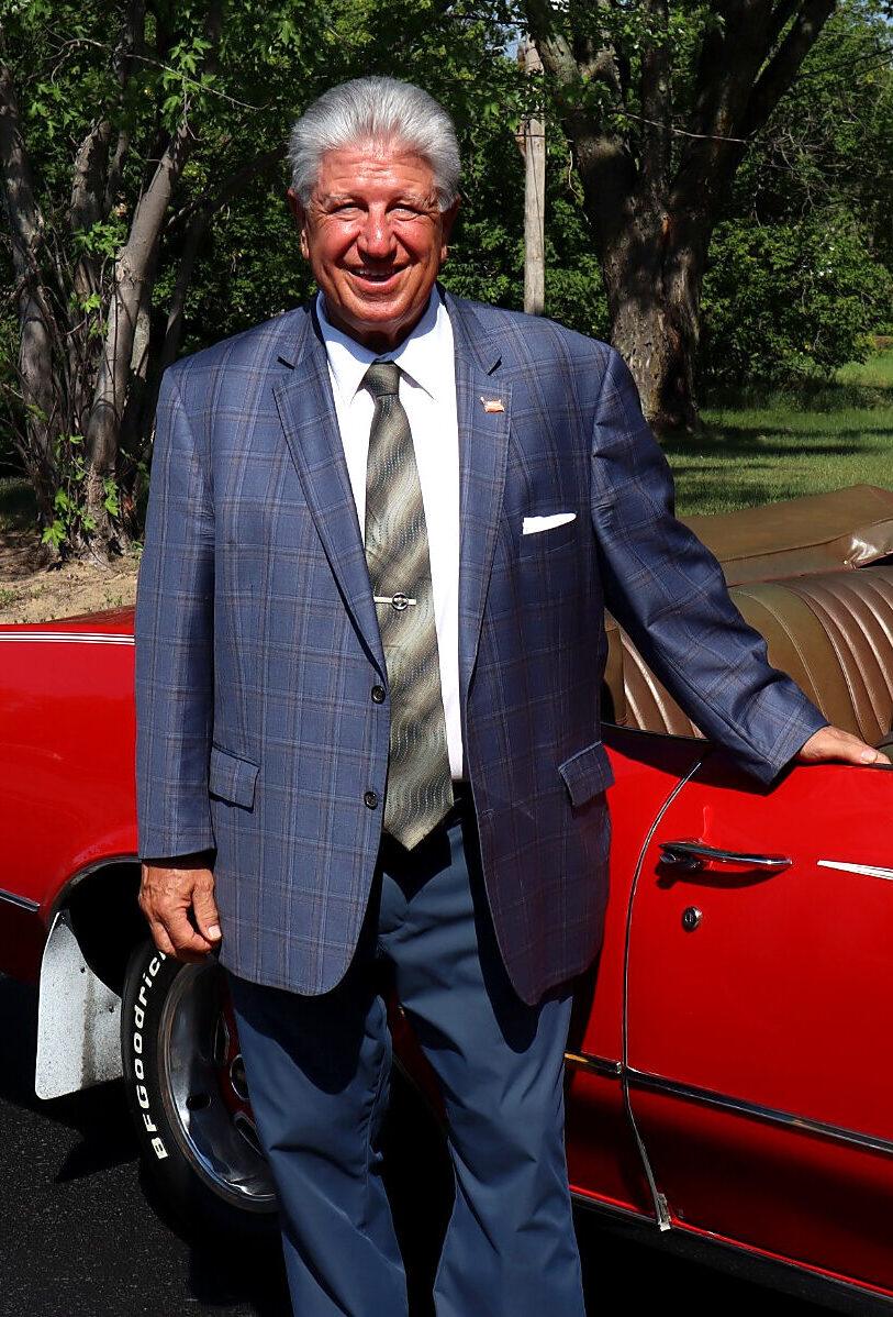 A smiling man in a plaid blazer and tie stands beside a red convertible car on a sunny day, exuding the confidence of a Business Home professional, with trees and greenery in the background.