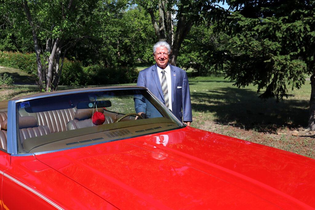 An older man in a suit stands outdoors next to a shiny red convertible car on a sunny day, surrounded by trees and greenery, looking ready to breakdance as he enjoys his success outside his business home.