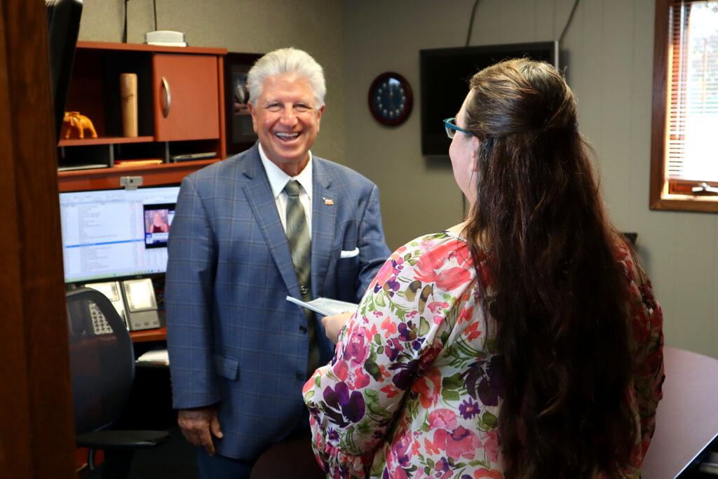 A man in a blue suit smiles while talking to a woman in a floral blouse inside a business home office with computers, shelves, and a window with blinds.