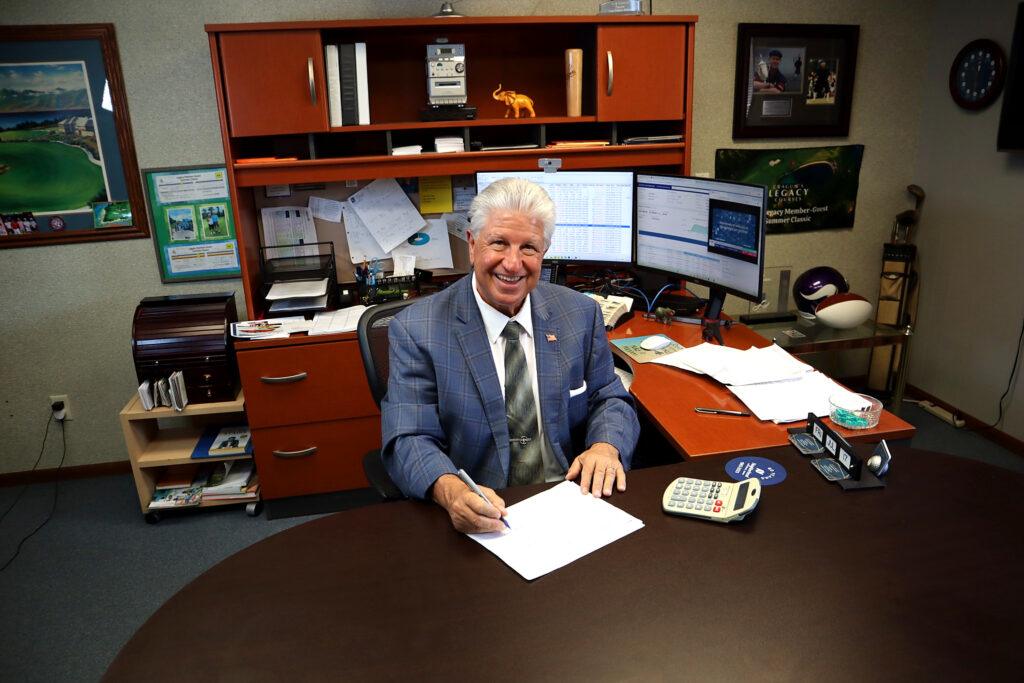 Smiling man in a suit sits at a desk in a Business Home office, holding a pen over papers. Behind him are shelves, multiple monitors, paperwork, and desk accessories. The office has a professional yet personalized appearance.