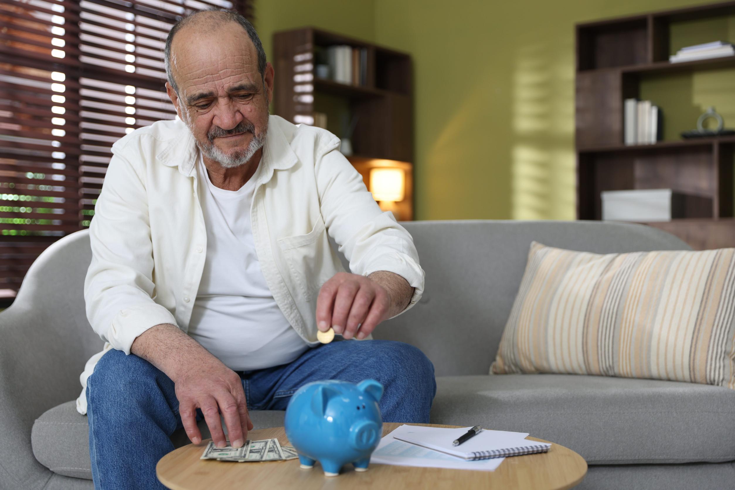 An older man sits on a sofa, placing a coin into a blue piggy bank on a table—a nostalgic symbol of retirement savings. Dollar bills, a notebook, and a pen are on the table, hinting at careful retirement planning. The room has warm lighting and shelves in the background.